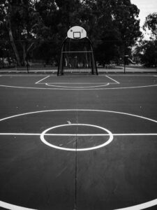 A minimalist black-and-white photograph of a deserted outdoor basketball court, hoop standing against an empty sky, no players present.