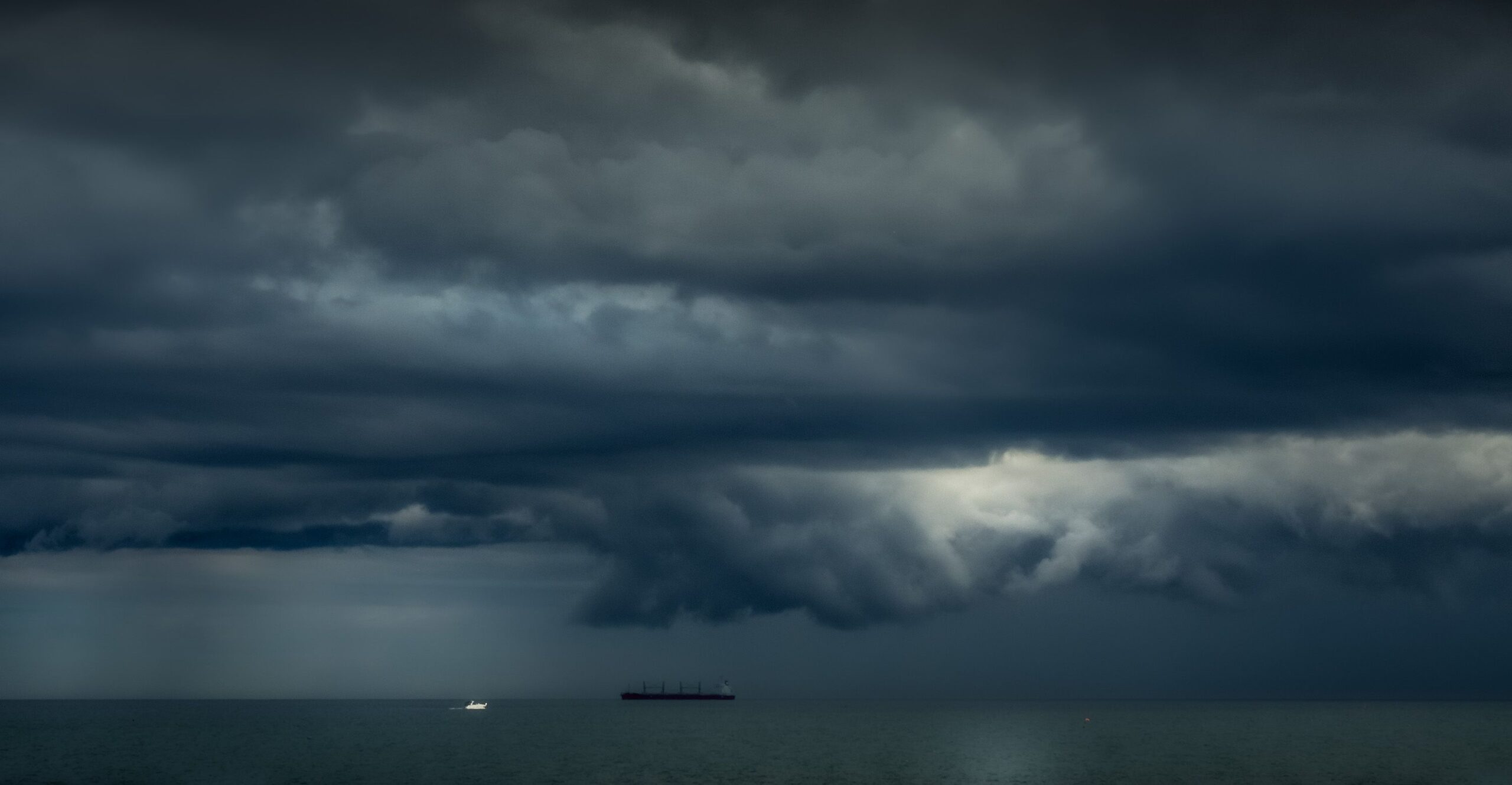 Dark storm clouds hanging low over a wide, open ocean, with faint silhouettes of ships on the distant horizon